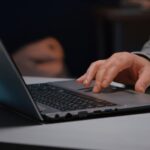 imagem de mão masculina digitando em um notebook que está em uma mesa branca